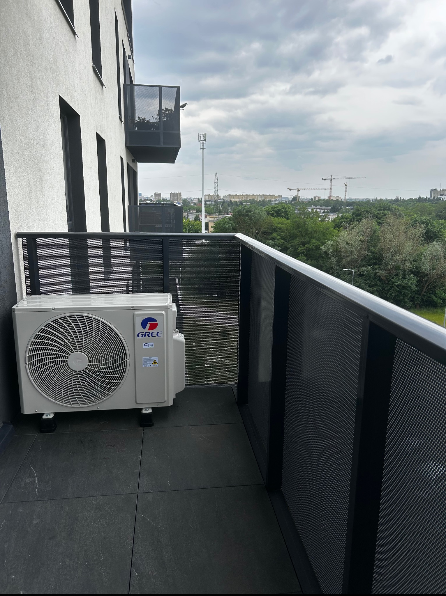 Klimatyzator Gree na balkonie nowoczesnego budynku. Widok na miasto z żurawiami budowlanymi w tle. Szare płytki na podłodze, ażurowa balustrada balkonu.
