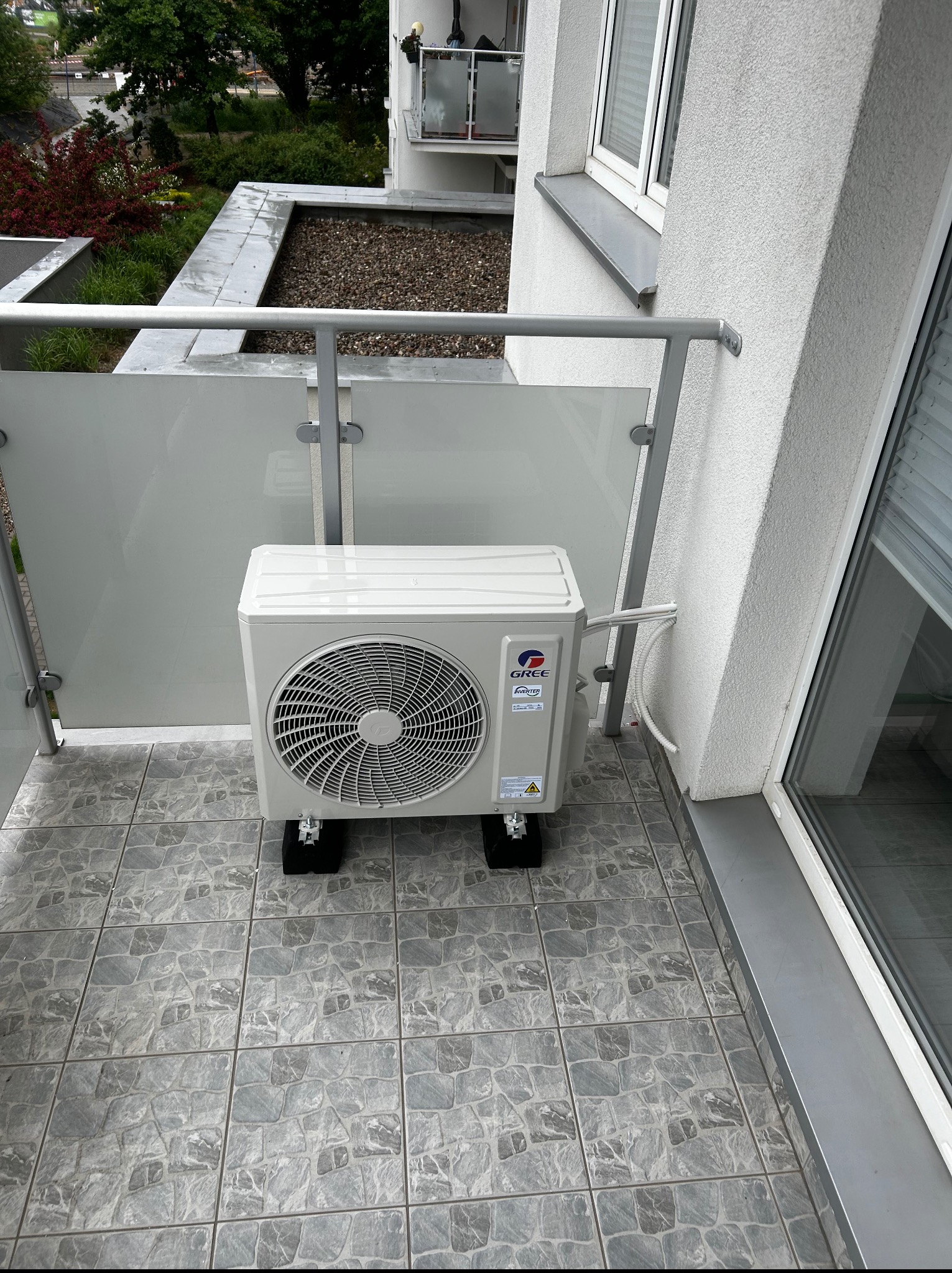 Biała jednostka zewnętrzna klimatyzacji Gree Inverter na balkonie z szarymi płytkami i przeszkloną balustradą. Widoczne przewody prowadzące do ściany budynku.