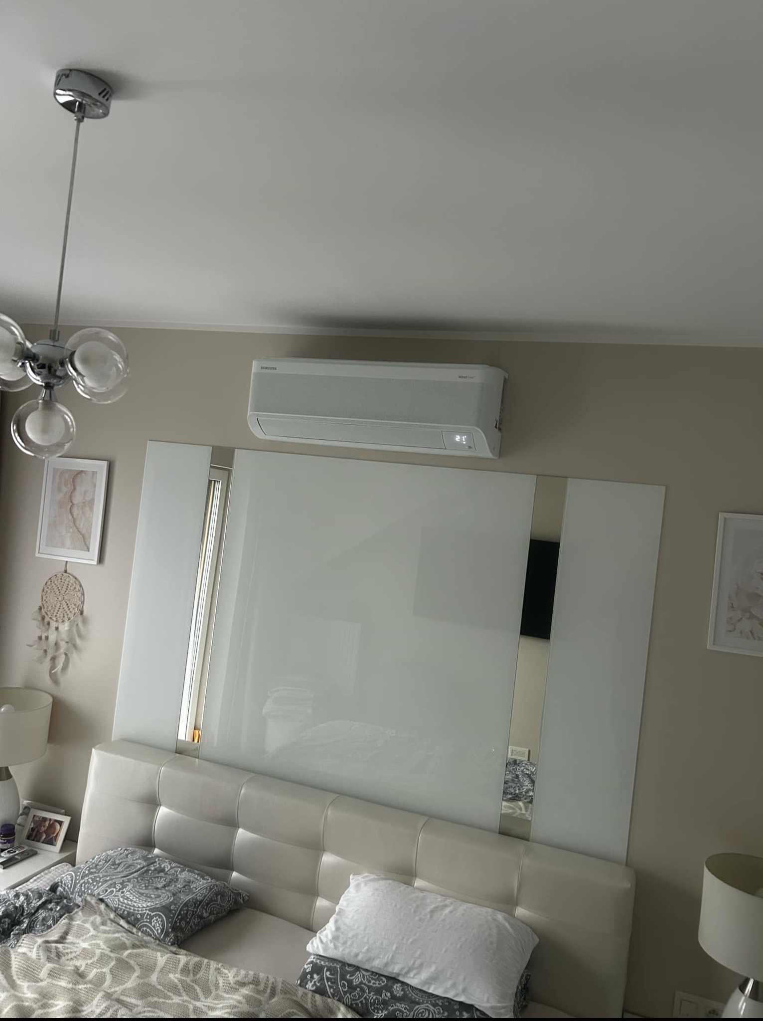 Biała klimatyzacja Samsung zamontowana nad łóżkiem w sypialni. Widoczny fragment lampy sufitowej i dekoracje ścienne. Minimalistyczny design wnętrza.