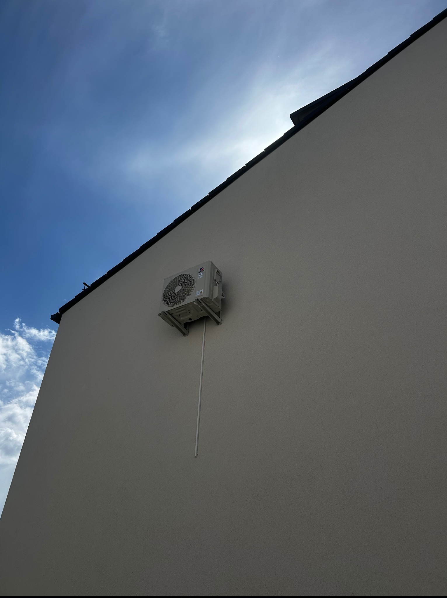 Zewnętrzna jednostka klimatyzacji zamontowana na elewacji budynku, widoczna z dołu pod kątem. Błękitne niebo z chmurami w tle. Biała rurka odprowadzająca skropliny.