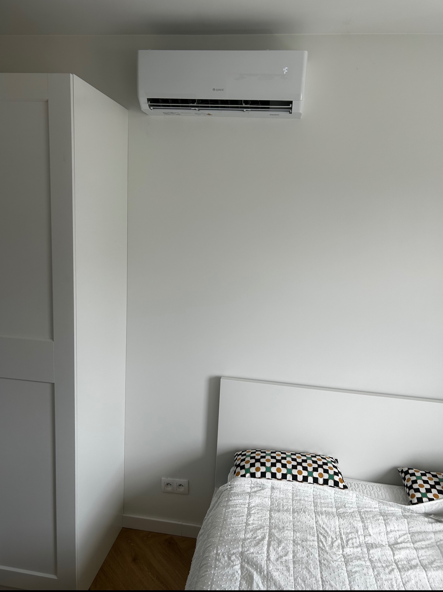Biały klimatyzator Gree zamontowany nad łóżkiem w minimalistycznej sypialni. Widoczne gniazdka elektryczne, biała szafa w tle.