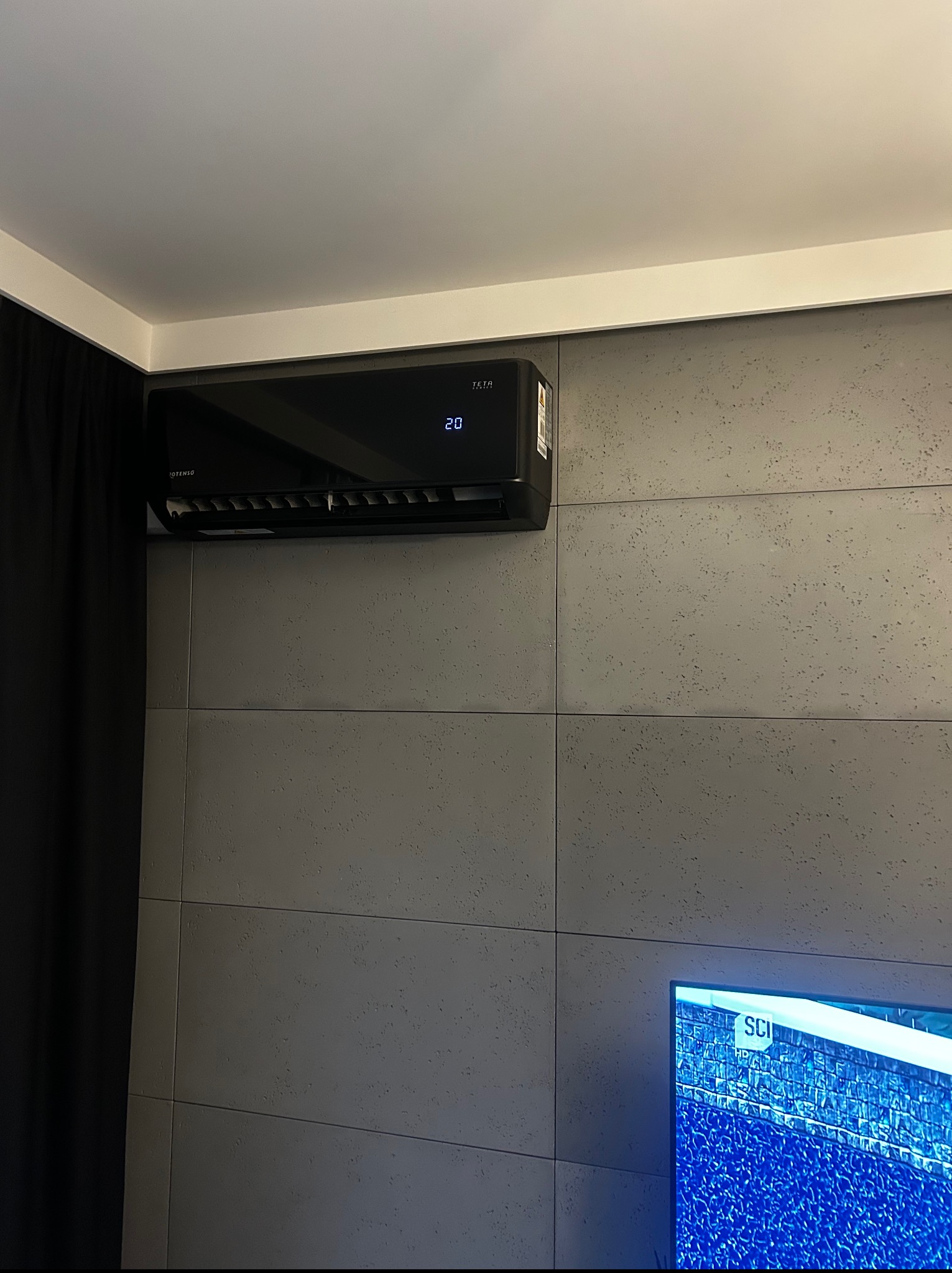 Czarna klimatyzacja ścienna z wyświetlaczem temperatury (20°C) na szarej ścianie z dekoracyjnymi panelami, obok czarna zasłona i fragment telewizora.
