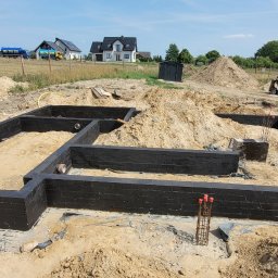 BETKO Roboty ogólnobudowlane - Czarne, murowane fundamenty domu na tle wykopu i sterty ziemi. Widoczne wystające pręty zbrojeniowe i elementy instalacji. W tle domy jednorodzinne i błękitne niebo.