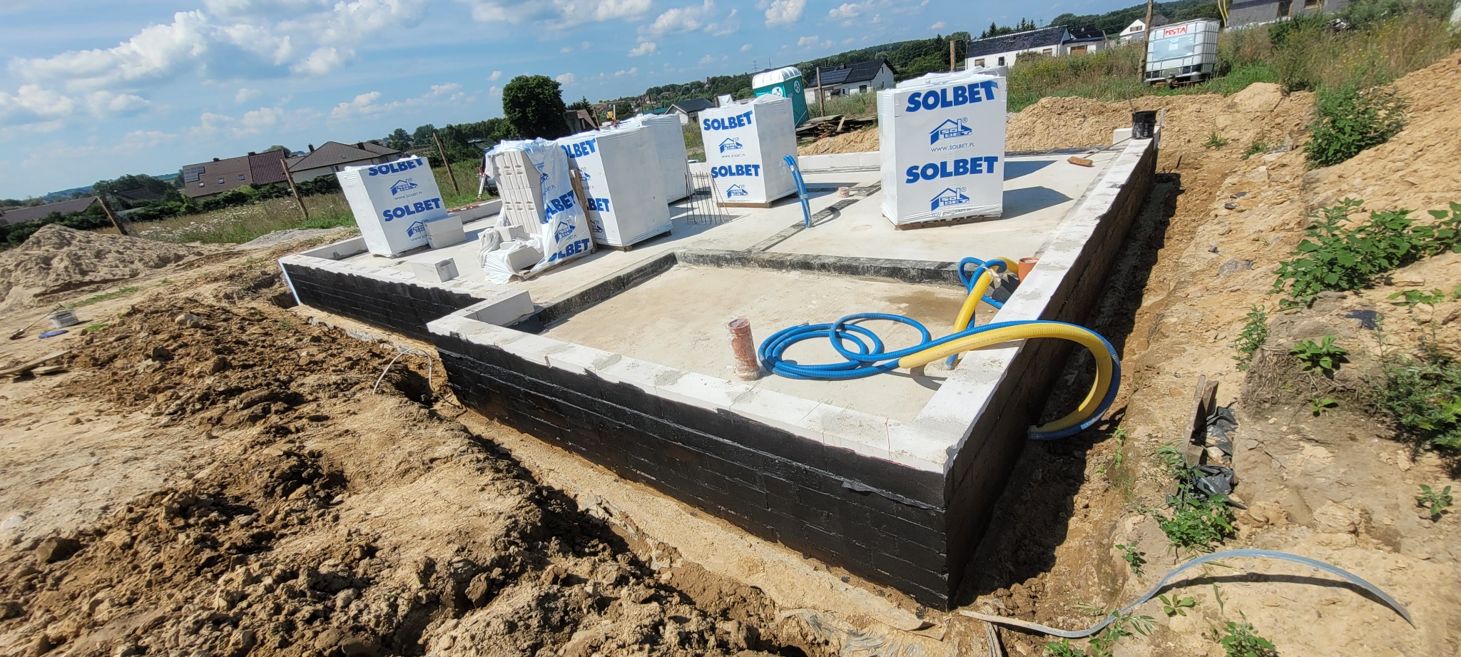 Fundamenty domu w trakcie budowy: ściany z izolacją przeciwwilgociową, bloczki Solbet, węże doprowadzające wodę. Widok z góry na plac budowy.