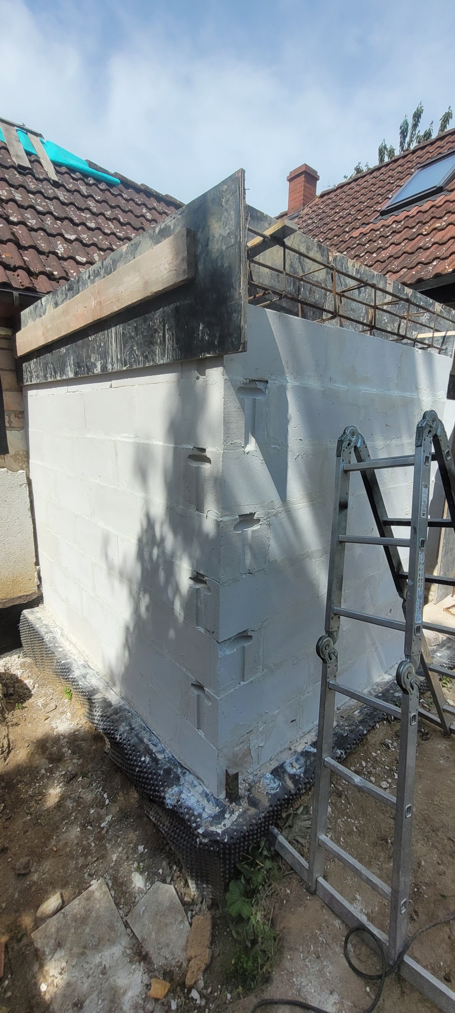 Wzniesiona ściana z białych bloczków, z widoczną izolacją fundamentów i szalunkiem. Metalowa drabina oparta o ścianę. Tło: dach budynku z dachówką.