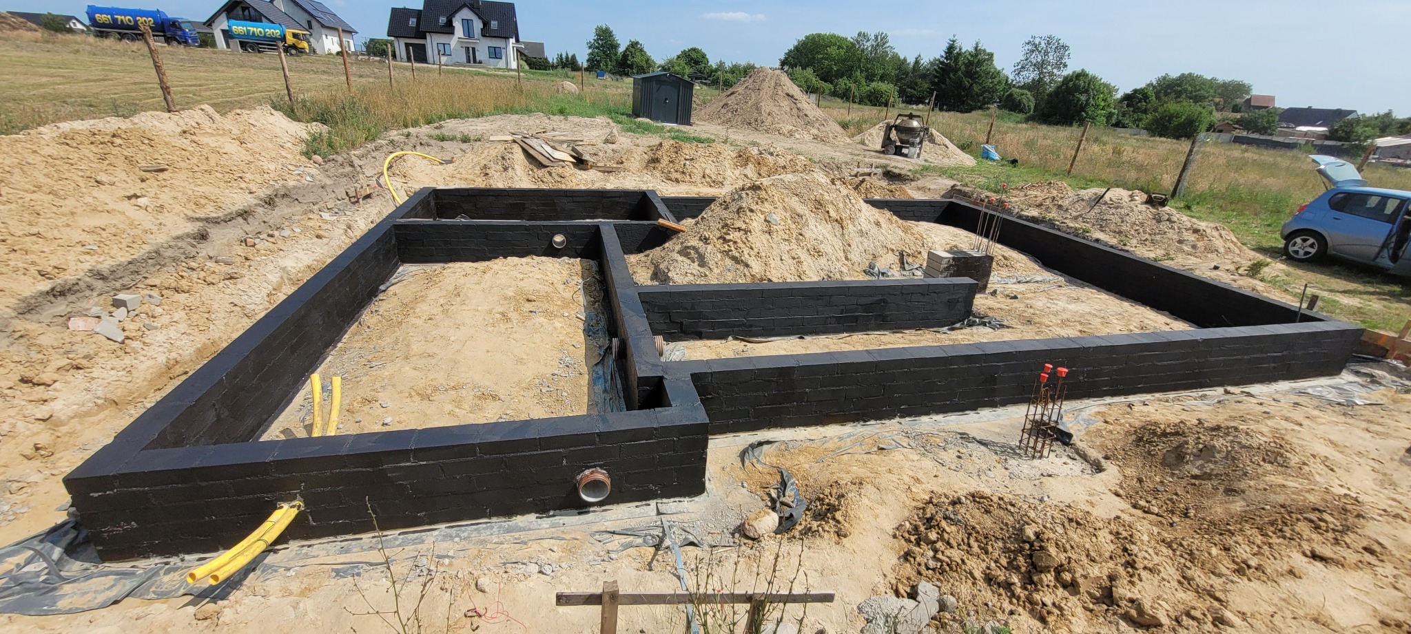 Czarno pomalowane, murowane ściany fundamentowe w wykopie, z widocznymi rurami i zbrojeniami. W tle dom w budowie i hałdy ziemi.