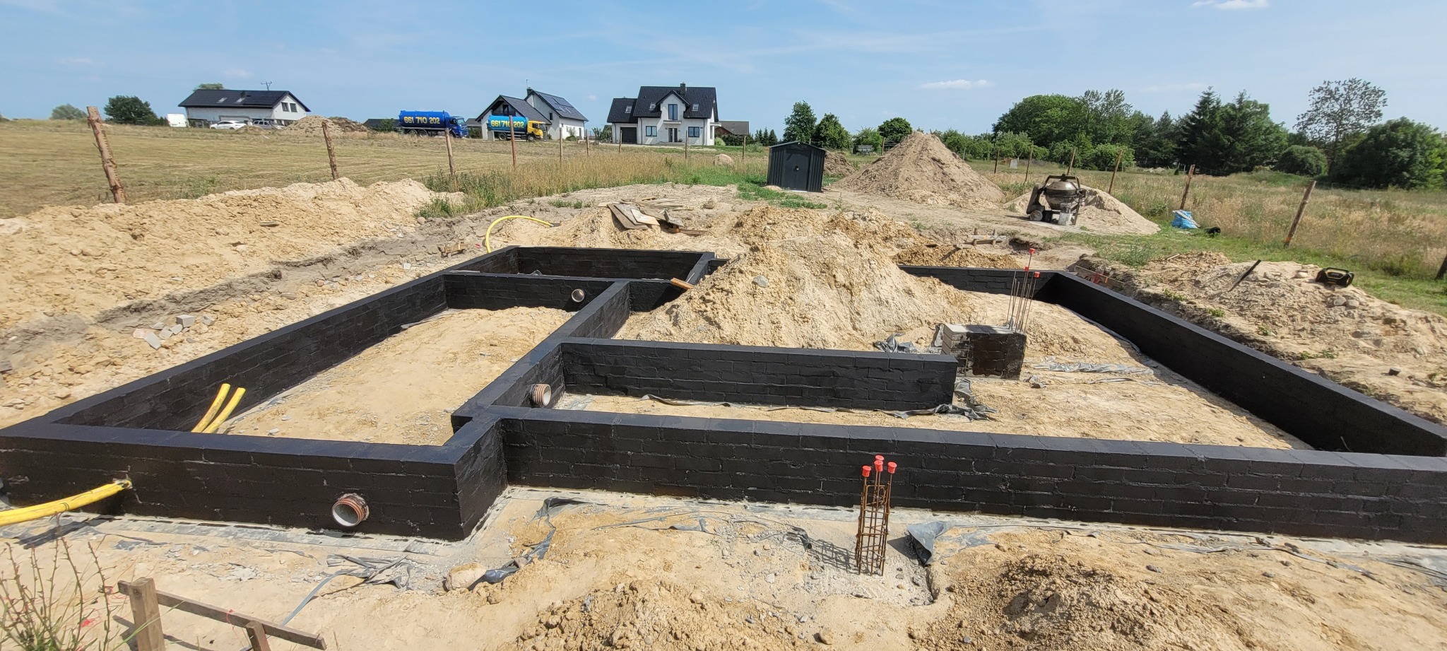 Czarne, murowane fundamenty domu na tle wykopu i sterty ziemi. Widoczne wystające pręty zbrojeniowe i elementy instalacji. W tle domy jednorodzinne i błękitne niebo.