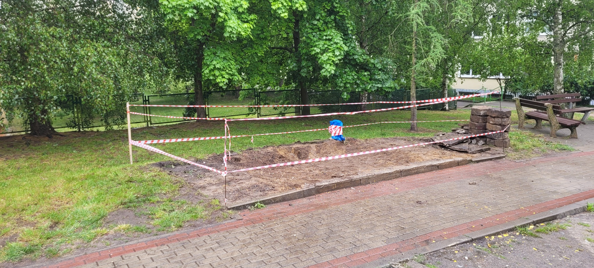 Teren po wykopie zabezpieczony taśmą ostrzegawczą na trawniku miejskim, obok stos kamiennych płyt i ławka parkowa.