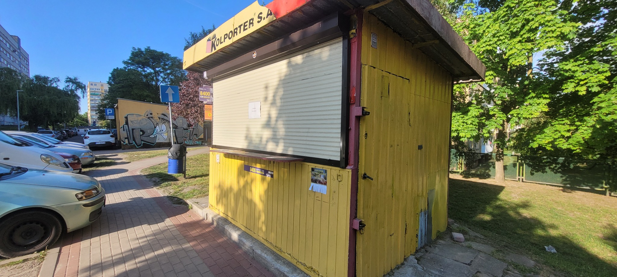 Żółty kiosk Kolporter S.A. z zasuwaną roletą, widoczny z boku na tle chodnika i zieleni. W tle budynki mieszkalne i zaparkowane samochody.