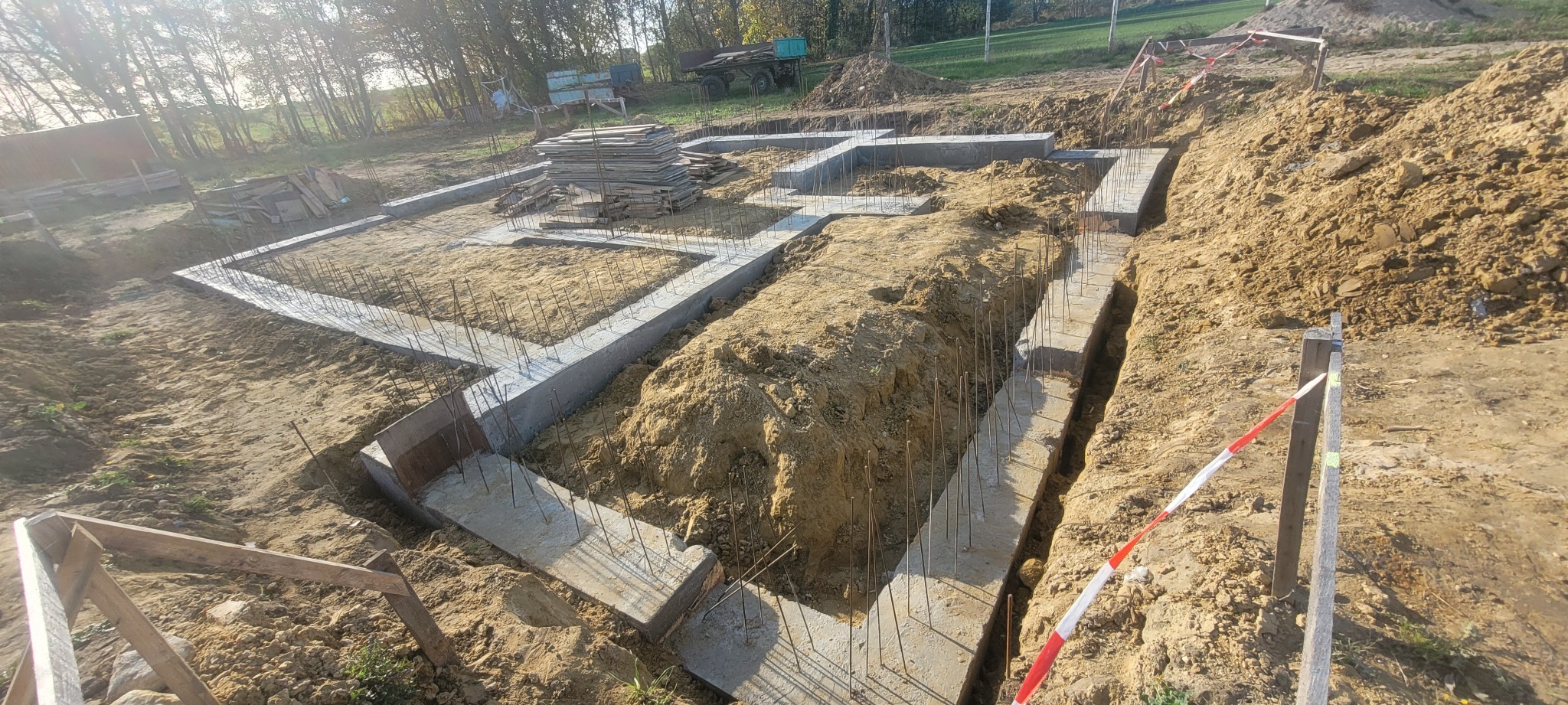 Zalane fundamenty z wystającymi prętami zbrojeniowymi, widok z góry na wykop, w tle drzewa i sprzęt budowlany. Etap prac fundamentowych.