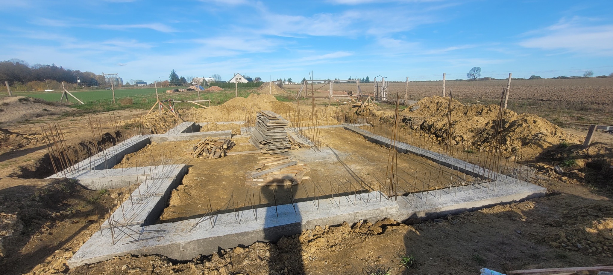 Zalane fundamenty domu jednorodzinnego z wystającymi prętami zbrojeniowymi, stos desek w wykopie, ziemia, błękitne niebo w tle. Widok z góry na plac budowy.
