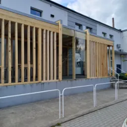 Nowoczesna drewniana konstrukcja przed budynkiem z przeszklonym wejściem, widok z zewnątrz, szara elewacja i białe barierki. Jasne drewno i minimalistyczny design.
