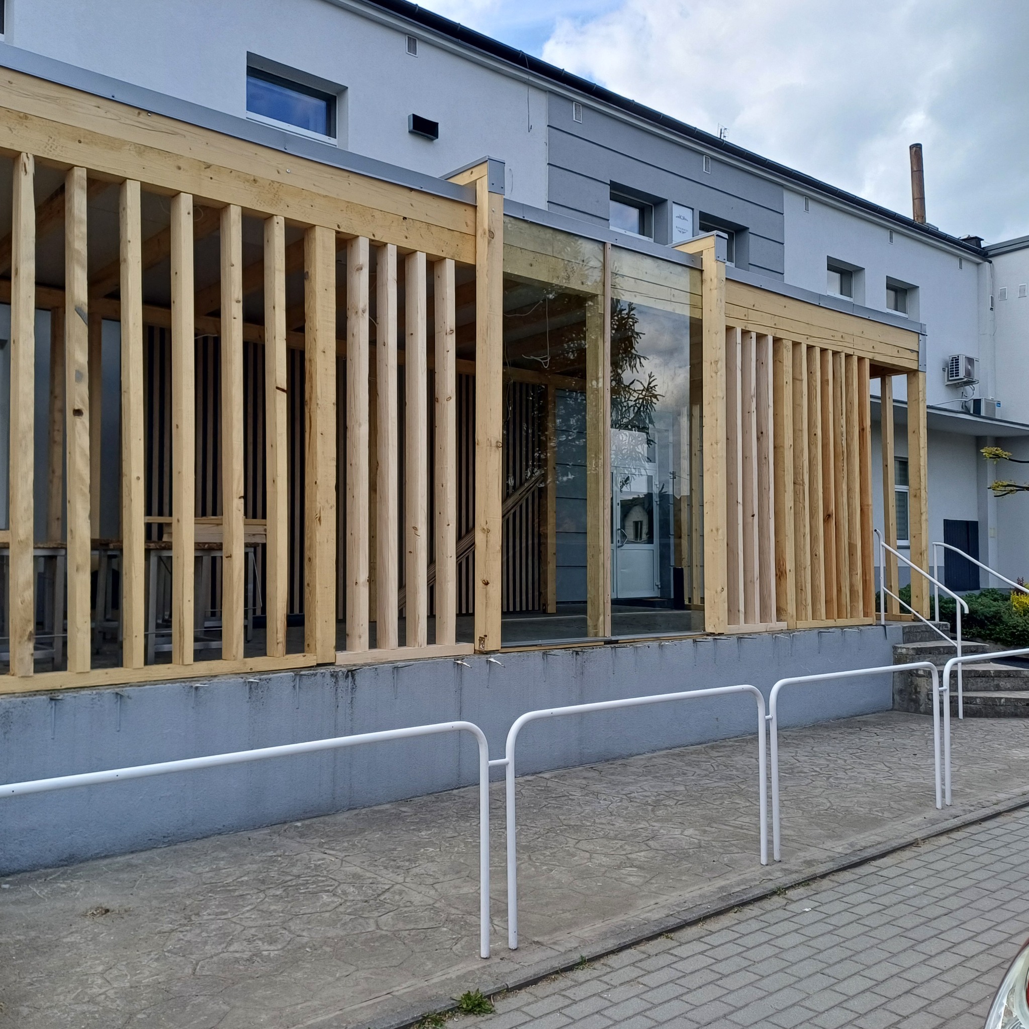 Nowoczesna drewniana konstrukcja przed budynkiem z przeszklonym wejściem, widok z zewnątrz, szara elewacja i białe barierki. Jasne drewno i minimalistyczny design.