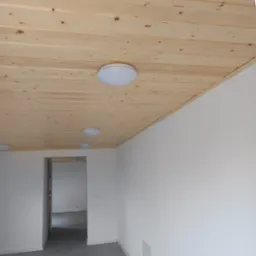 Jasne, drewniane panele sufitowe z trzema okrągłymi lampami LED w nowym, pomalowanym na biało pomieszczeniu z szarymi płytkami podłogowymi i widocznym przejściem do kolejnego pokoju.