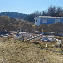 Wykopane fundamenty z częściowo wymurowanymi ścianami z bloczków betonowych. Na pierwszym planie palety z bloczkami, w tle ziemia i mały niebieski domek.