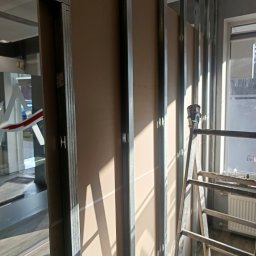 Bartosz Piszczyk - Montaż ścianki działowej z płyt karton-gips na metalowym stelażu, widoczna drabina i okno z grzejnikiem w tle. Częściowo widoczne pomieszczenie za ścianką.