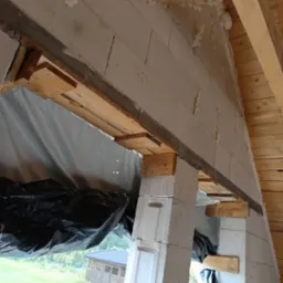 Wznoszenie ściany z betonu komórkowego Ytong z drewnianym szalunkiem okiennym, widoczne ocieplenie pianką montażową i drewniana więźba dachowa.