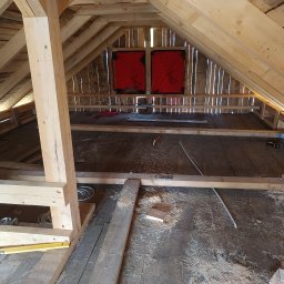 Bartosz Piszczyk - Poddasze w trakcie adaptacji: widoczne drewniane elementy konstrukcyjne, deski podłogowe pokryte wiórami, narzędzia i materiały budowlane. Dwa okna dachowe z czerwoną folią.