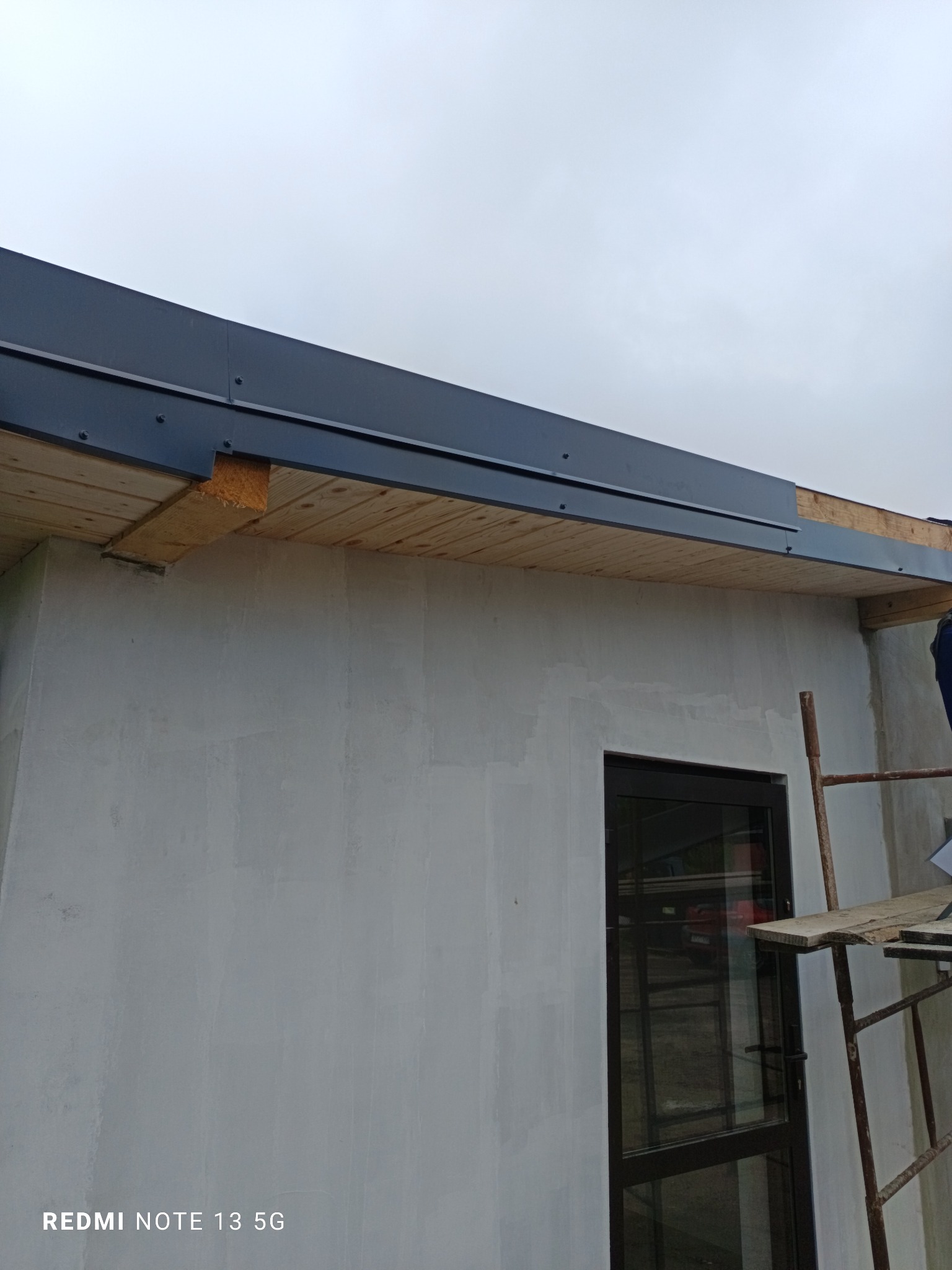 Wykończenie elewacji domu: szara ściana po tynkowaniu, widoczne drewniane elementy dachu i czarne wykończenie okapu, fragment rusztowania z boku.