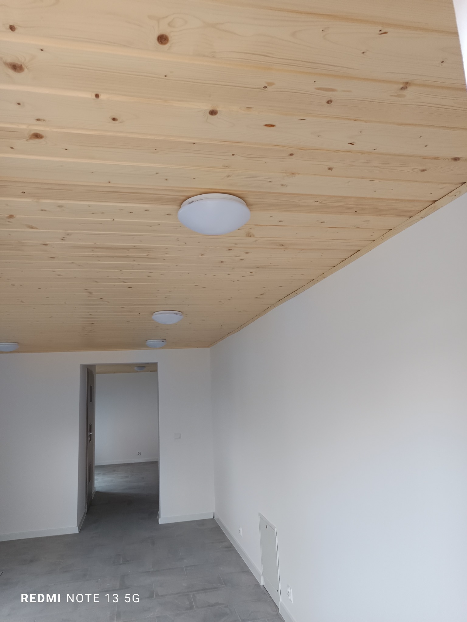 Jasne, drewniane panele sufitowe z trzema okrągłymi lampami LED w nowym, pomalowanym na biało pomieszczeniu z szarymi płytkami podłogowymi i widocznym przejściem do kolejnego pokoju.