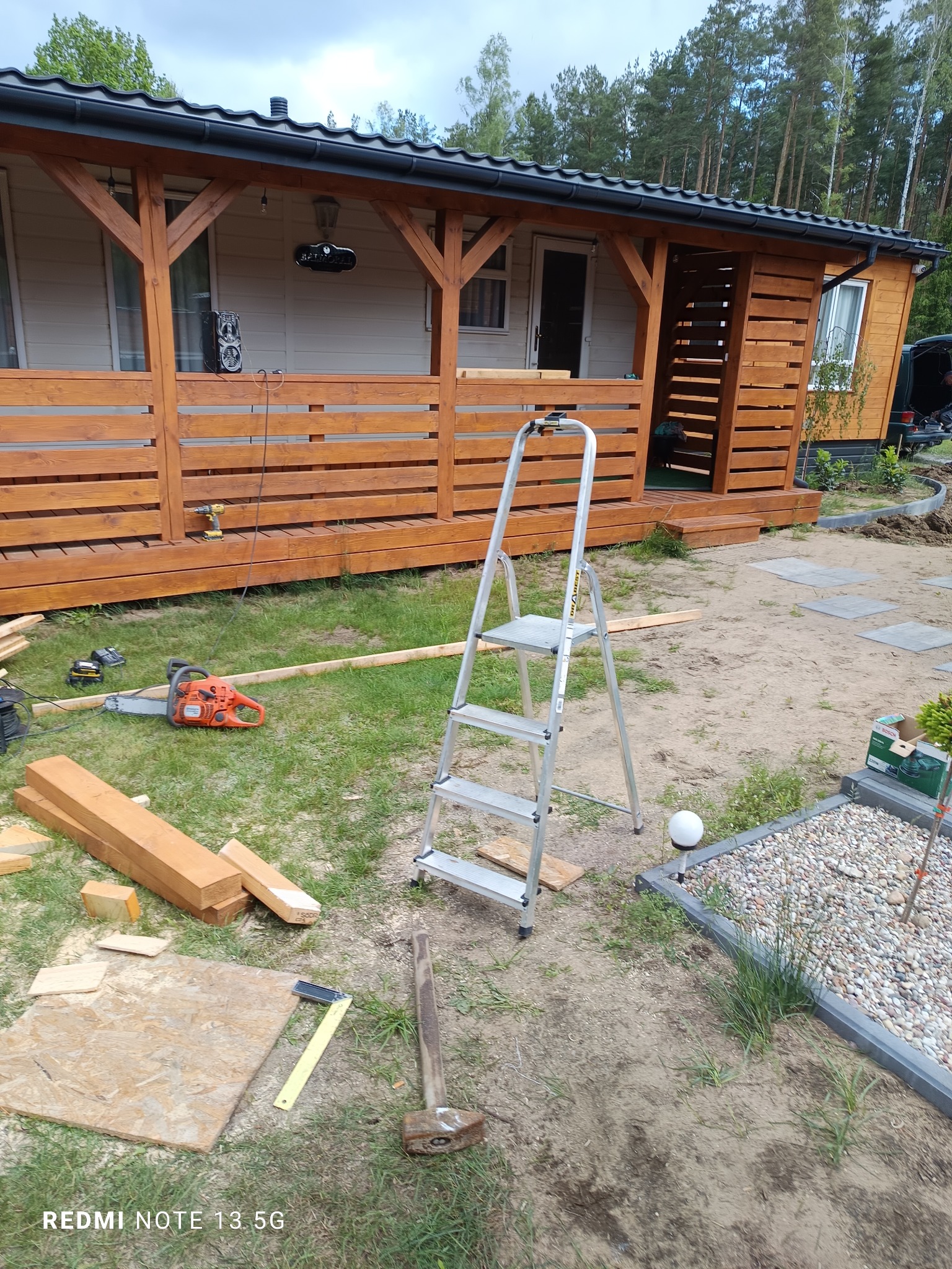 Drewniany domek letniskowy z zadaszonym tarasem w trakcie budowy, widoczne narzędzia: pilarka, wiertarka, drabina, młotek i elementy konstrukcyjne.