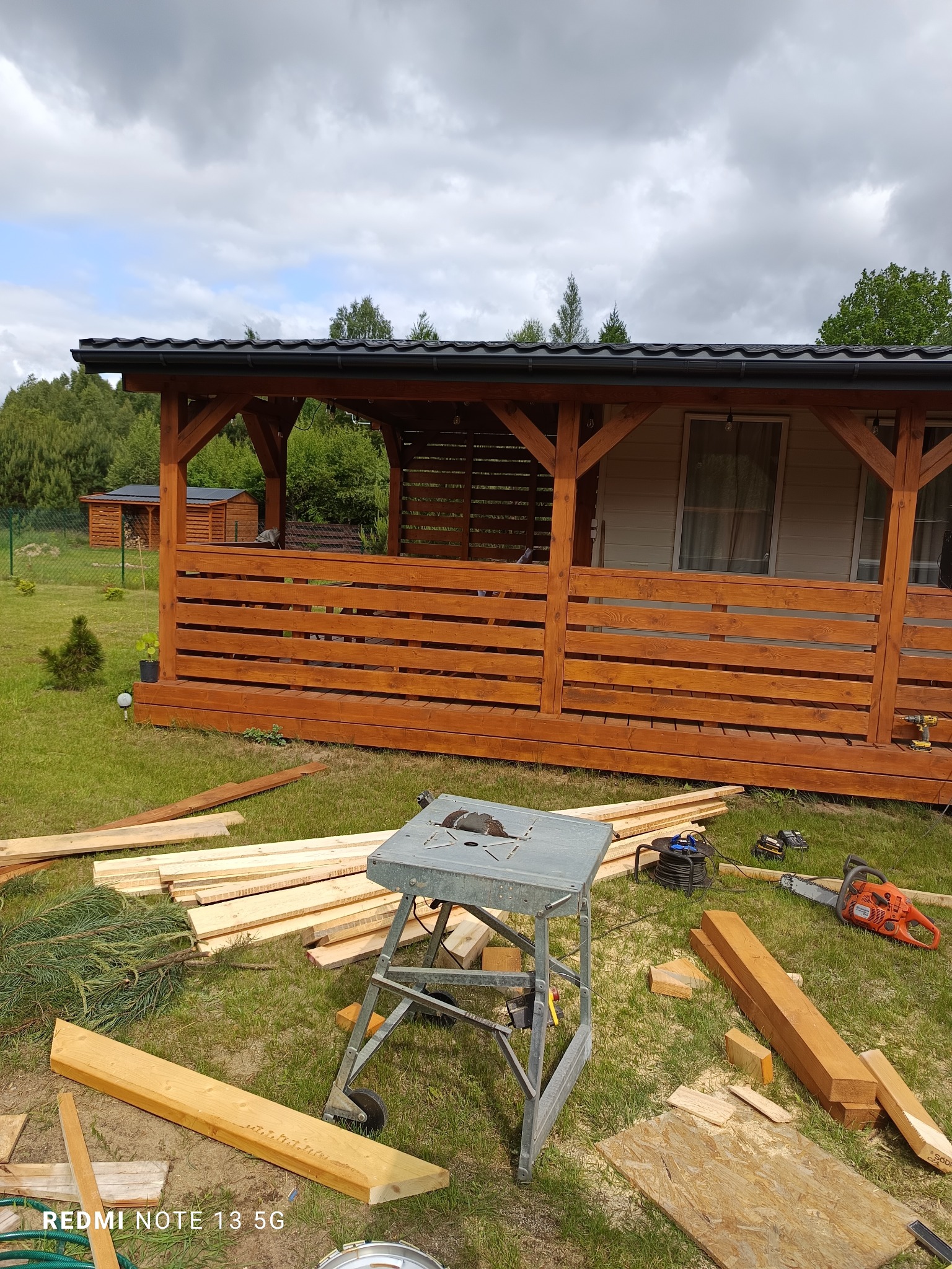 Drewniana altana z brązową balustradą w trakcie budowy, widoczne narzędzia i materiały na trawie, drzewa w tle. Kąt ujęcia z dołu.