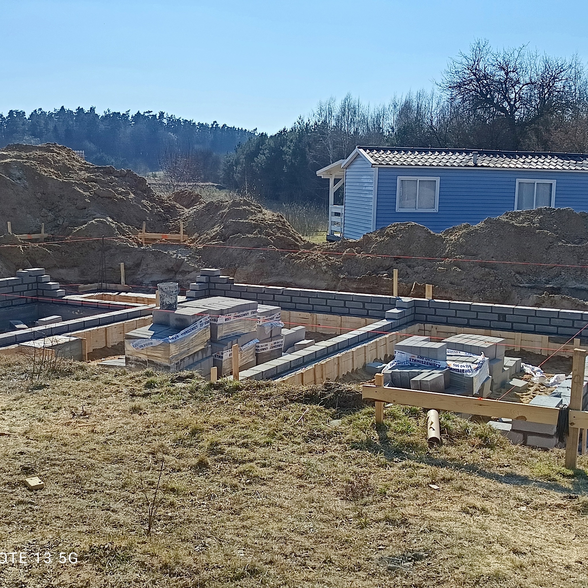 Wykopane fundamenty z częściowo wymurowanymi ścianami z bloczków betonowych. Na pierwszym planie palety z bloczkami, w tle ziemia i mały niebieski domek.