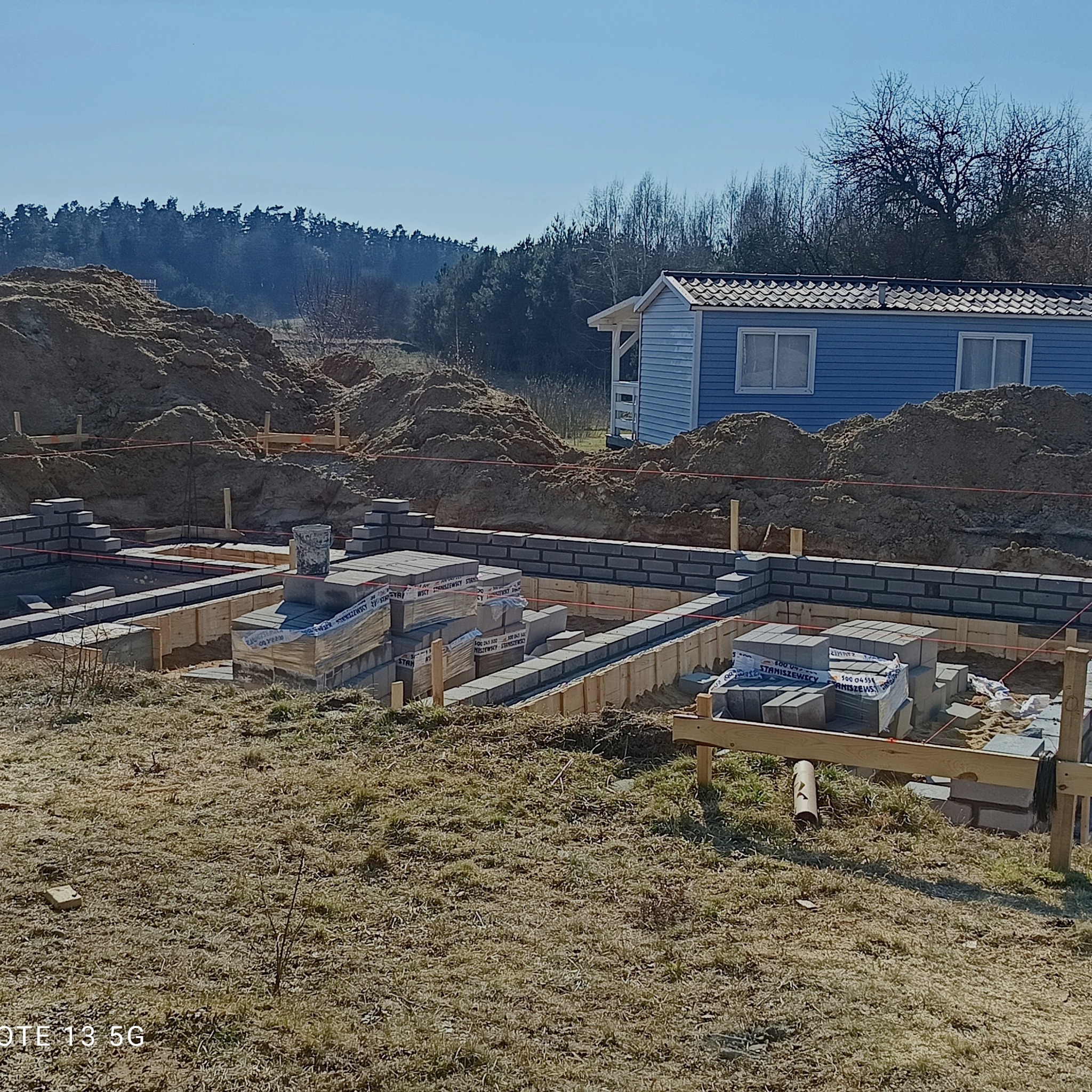 Budowa fundamentów z bloczków betonowych na działce z małym domkiem w tle. Widoczne drewniane szalunki i zapasy materiałów budowlanych.