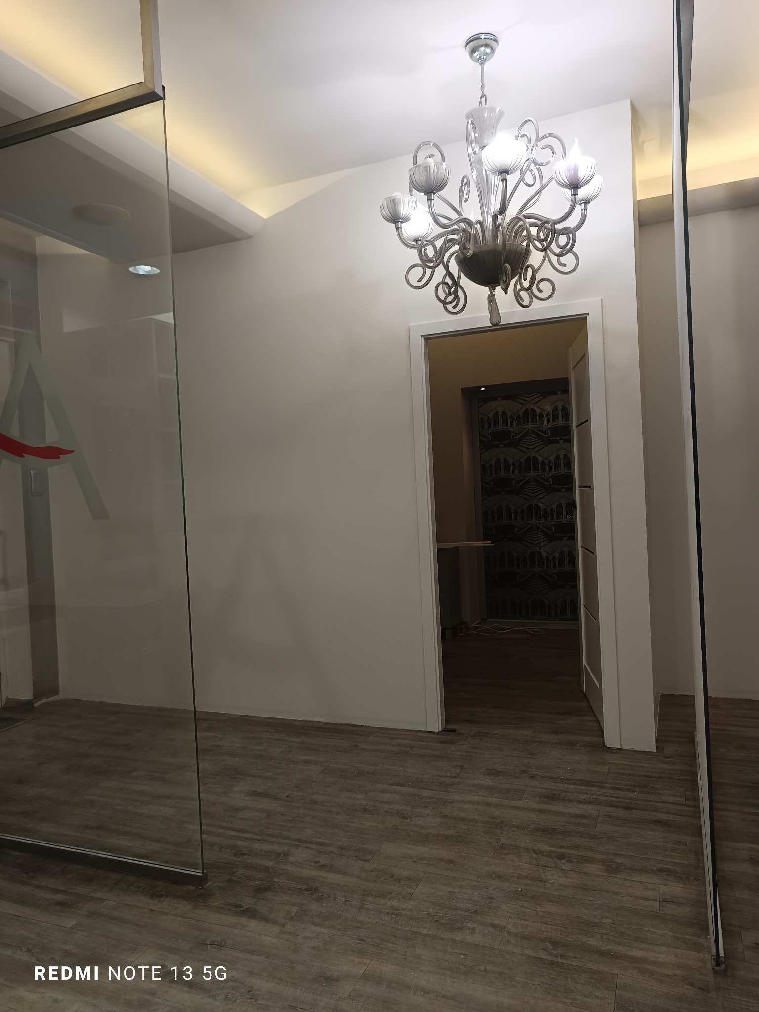 Jasne wnętrze z parkietem, widok na pokój z wzorzystą tapetą przez otwarte drzwi. Szklana ściana z logo firmy, elegancki żyrandol. Nowoczesny design.