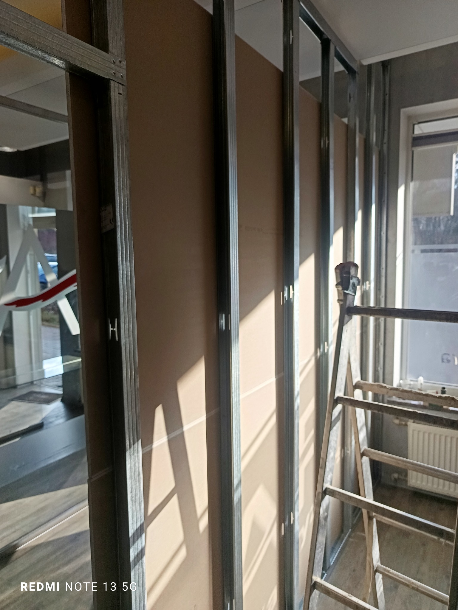 Montaż ścianki działowej z płyt karton-gips na metalowym stelażu, widoczna drabina i okno z grzejnikiem w tle. Częściowo widoczne pomieszczenie za ścianką.