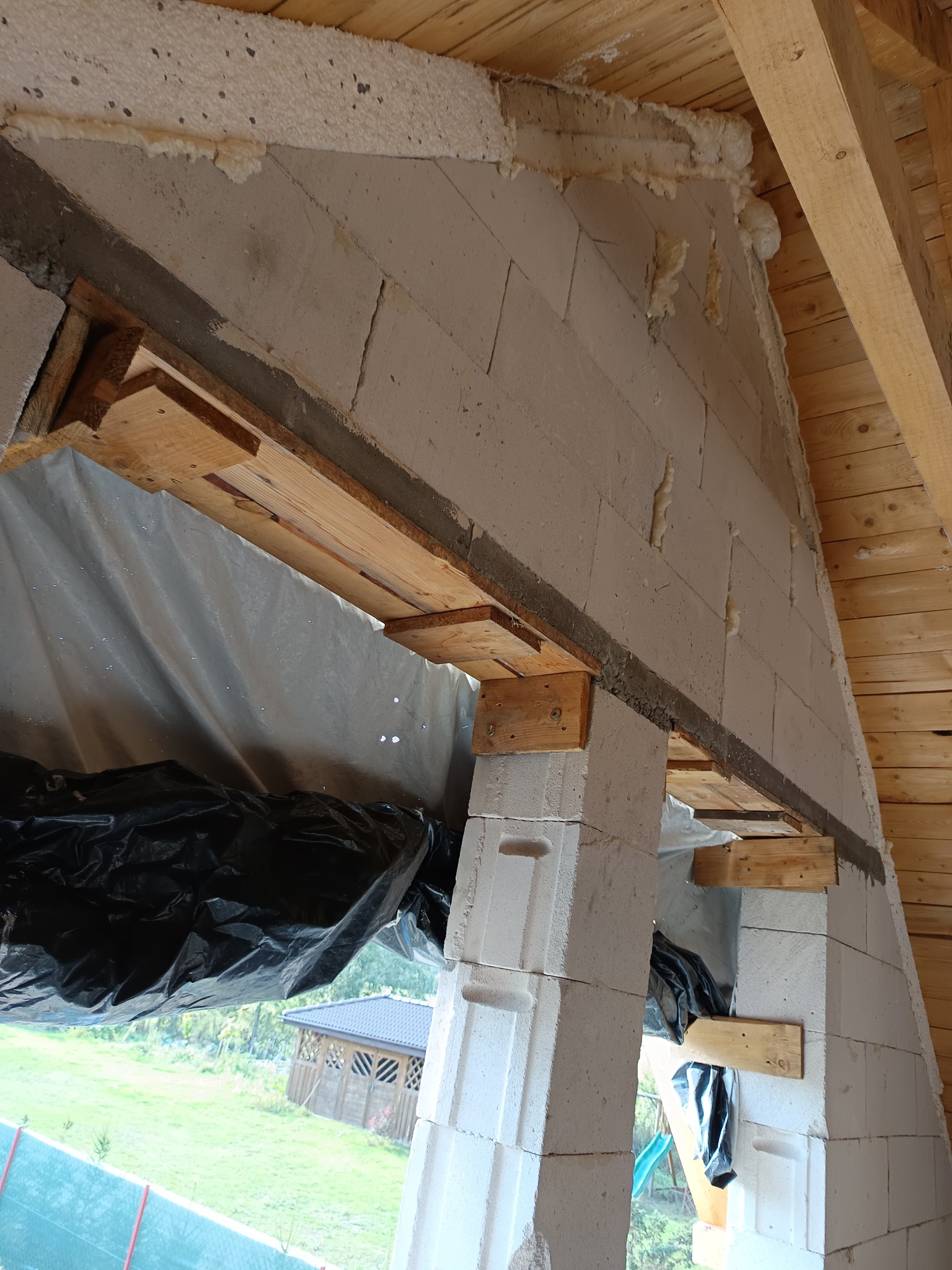 Wznoszenie ściany z betonu komórkowego Ytong z drewnianym szalunkiem okiennym, widoczne ocieplenie pianką montażową i drewniana więźba dachowa.