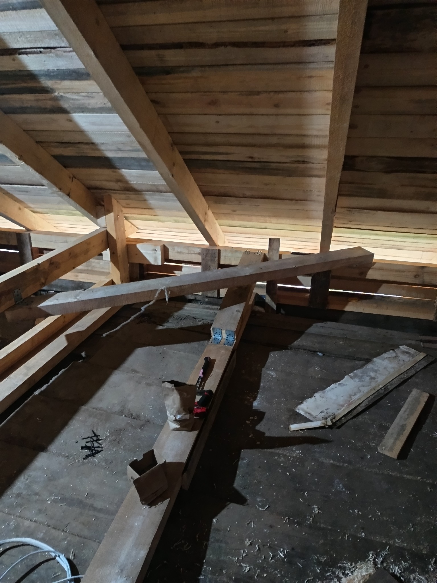 Wnętrze poddasza z drewnianą konstrukcją dachu w trakcie budowy. Widoczne deski, belki, narzędzia i kable. Surowe wykończenie, etap prac w toku.