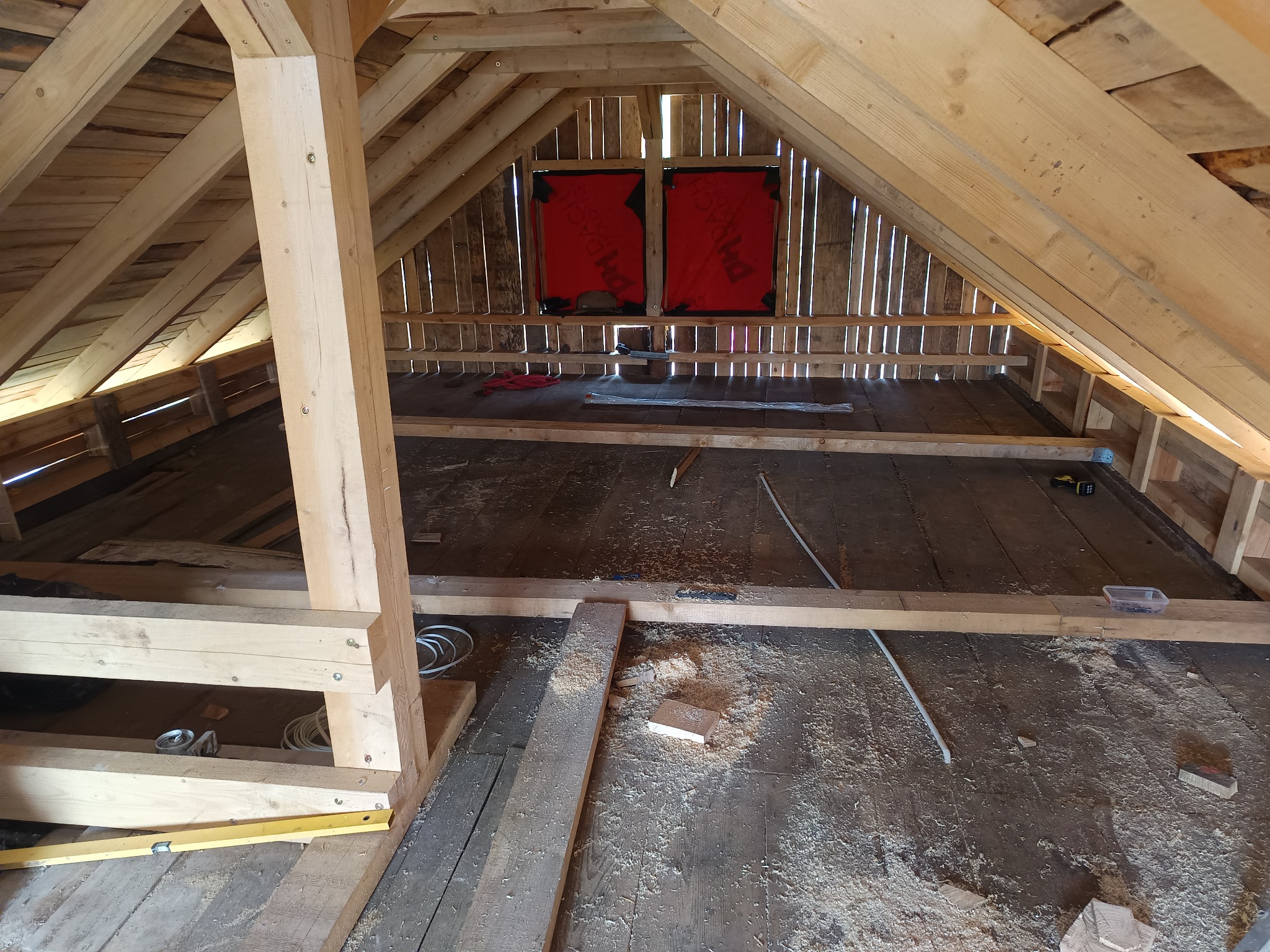 Poddasze w trakcie adaptacji: widoczne drewniane elementy konstrukcyjne, deski podłogowe pokryte wiórami, narzędzia i materiały budowlane. Dwa okna dachowe z czerwoną folią.