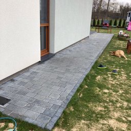 ECO CLEANING RENATA KOWZIEL - Świeżo ułożona, szara kostka brukowa przy białej elewacji domu. Na trawie leżą klapki i pies. Widoczne drzewa w tle.
