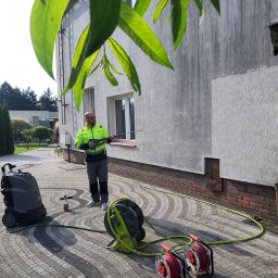 ECO CLEANING RENATA KOWZIEL - Pracownik w odblaskowej kurtce czyści elewację domu myjką ciśnieniową. Widoczny sprzęt, wąż i kostka brukowa. Ujęcie z dołu, z liśćmi drzewa w kadrze.