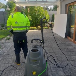 ECO CLEANING RENATA KOWZIEL - Pracownik w odblaskowej kurtce stoi tyłem obok myjki ciśnieniowej Kärcher na kostce brukowej. Widoczny fragment elewacji domu i zieleń w tle.