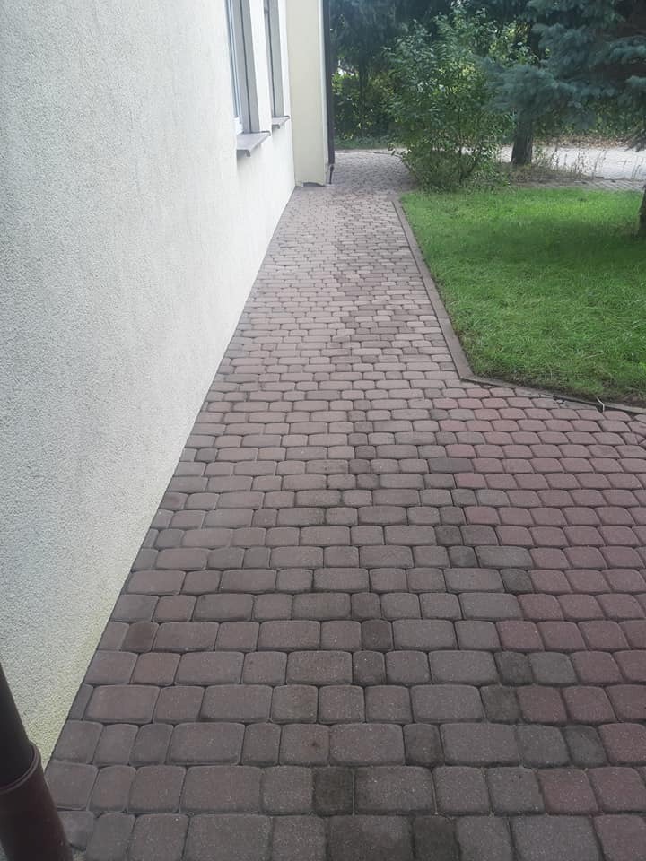 Czysta, brązowa kostka brukowa ułożona wzdłuż jasnej ściany budynku i trawnika. Widok z góry, ukazujący regularny wzór kostki i zadbane otoczenie.