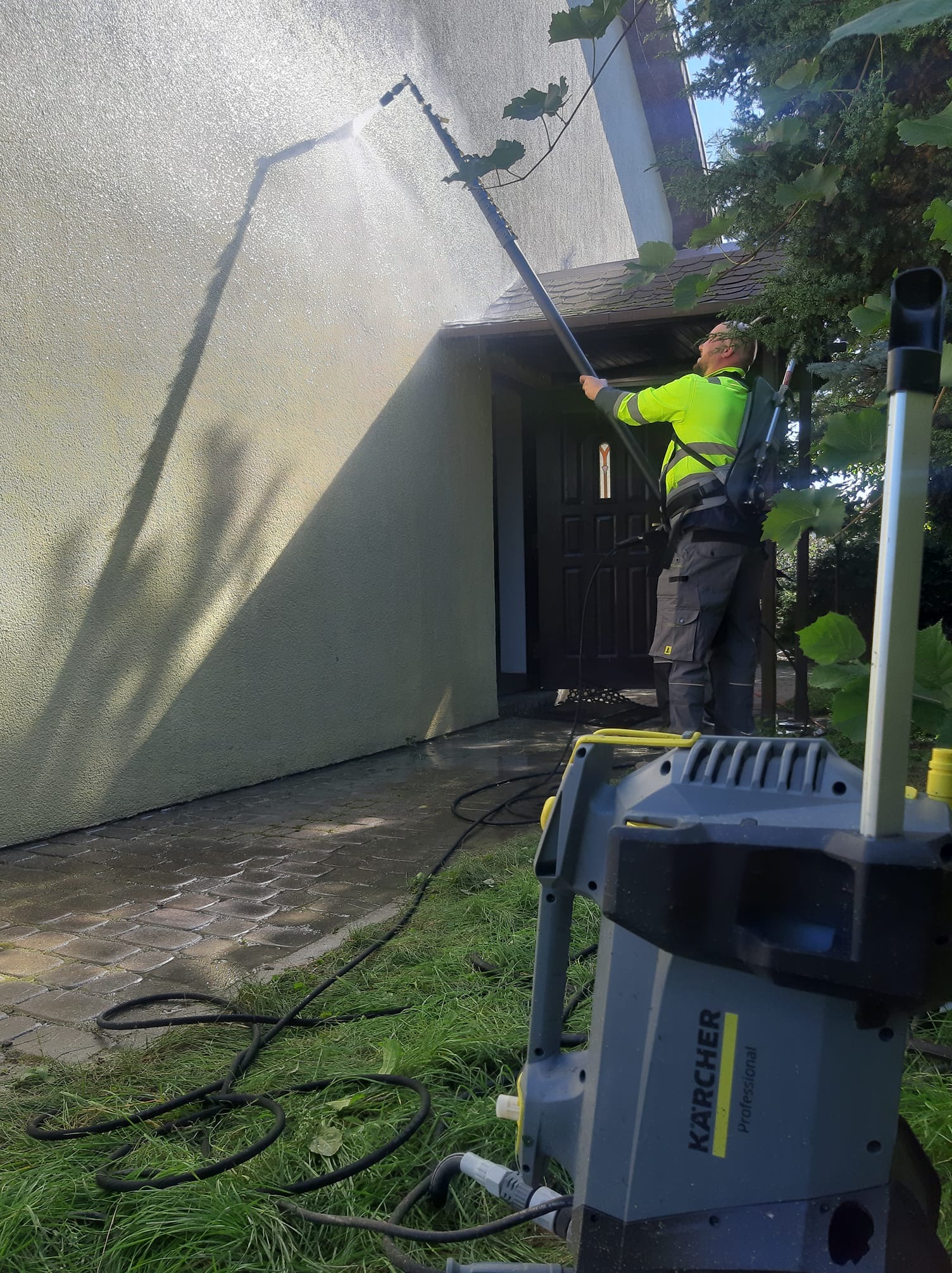 Pracownik w jaskrawej kamizelce czyści elewację domu za pomocą myjki ciśnieniowej Kärcher. Widoczny sprzęt i woda spływająca po ścianie. Ujęcie z dołu.