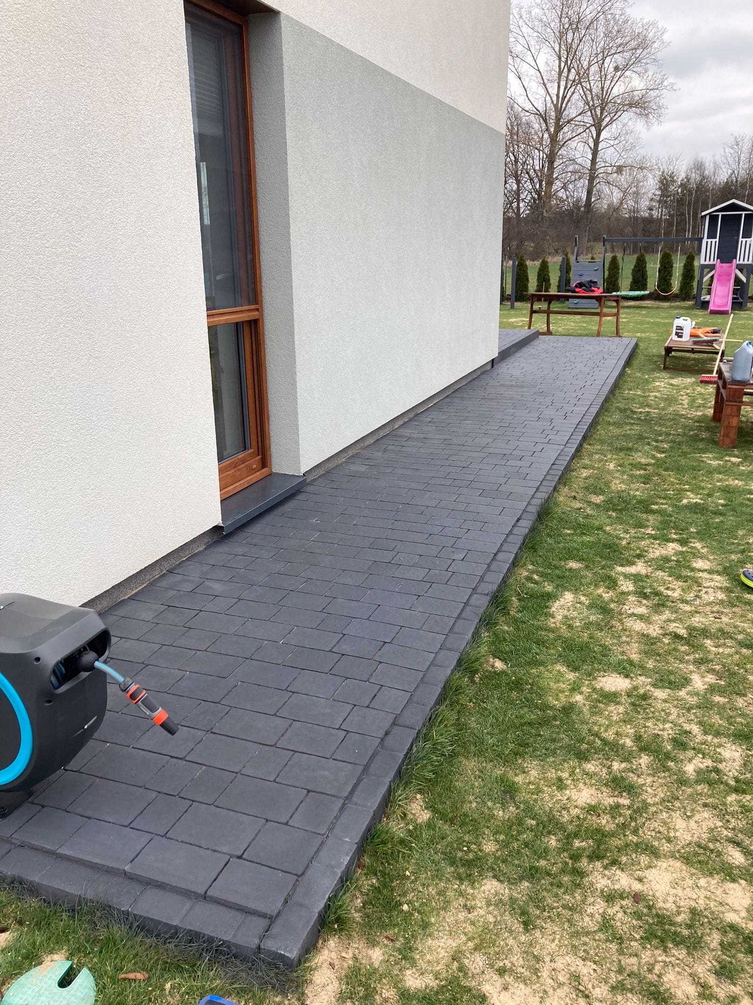 Ciemnoszara kostka brukowa prowadząca od elewacji domu do zielonego trawnika. Widoczny zwijak węża ogrodowego, okno i plac zabaw w tle.