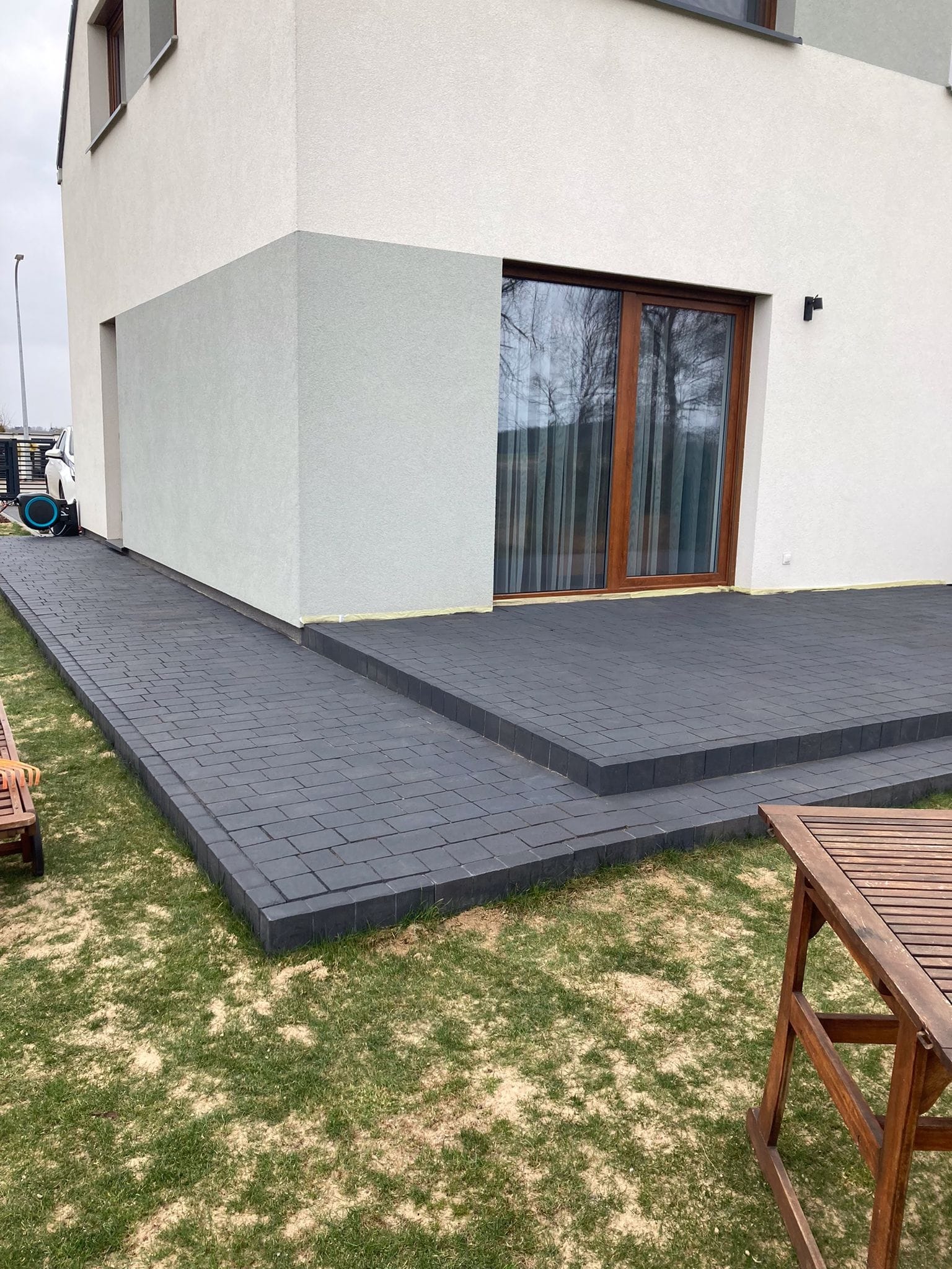Nowoczesny dom z ciemną kostką brukową tworzącą ścieżkę i taras. Widoczne schody z kostki, fragment drewnianego stołu i trawnik. Dom w jasnych barwach.