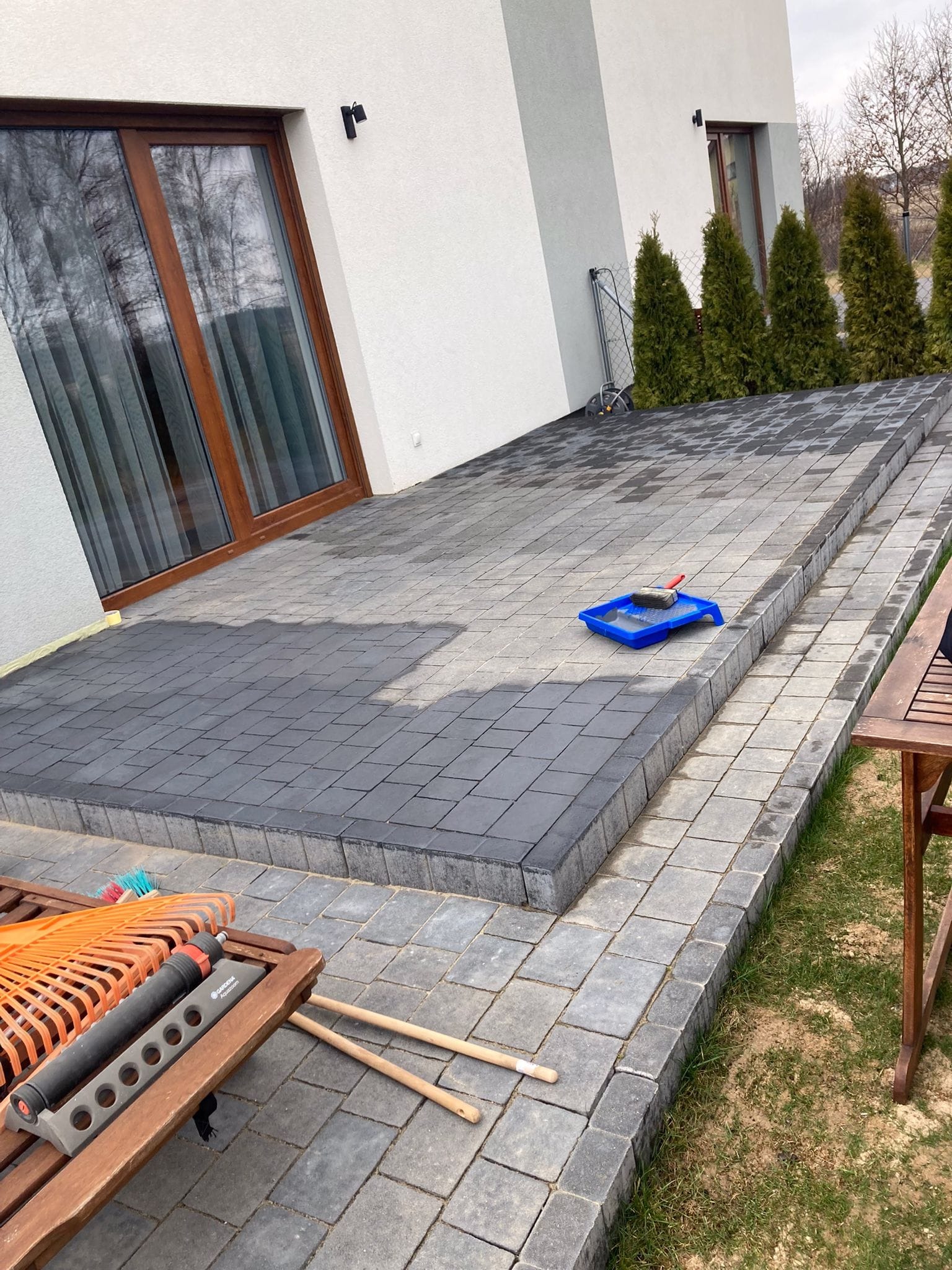 Częściowo wyczyszczona kostka brukowa na tarasie domu z widocznym narzędziem (kuweta z wałkiem). Widoczny kontrast między czystą a zabrudzoną powierzchnią.