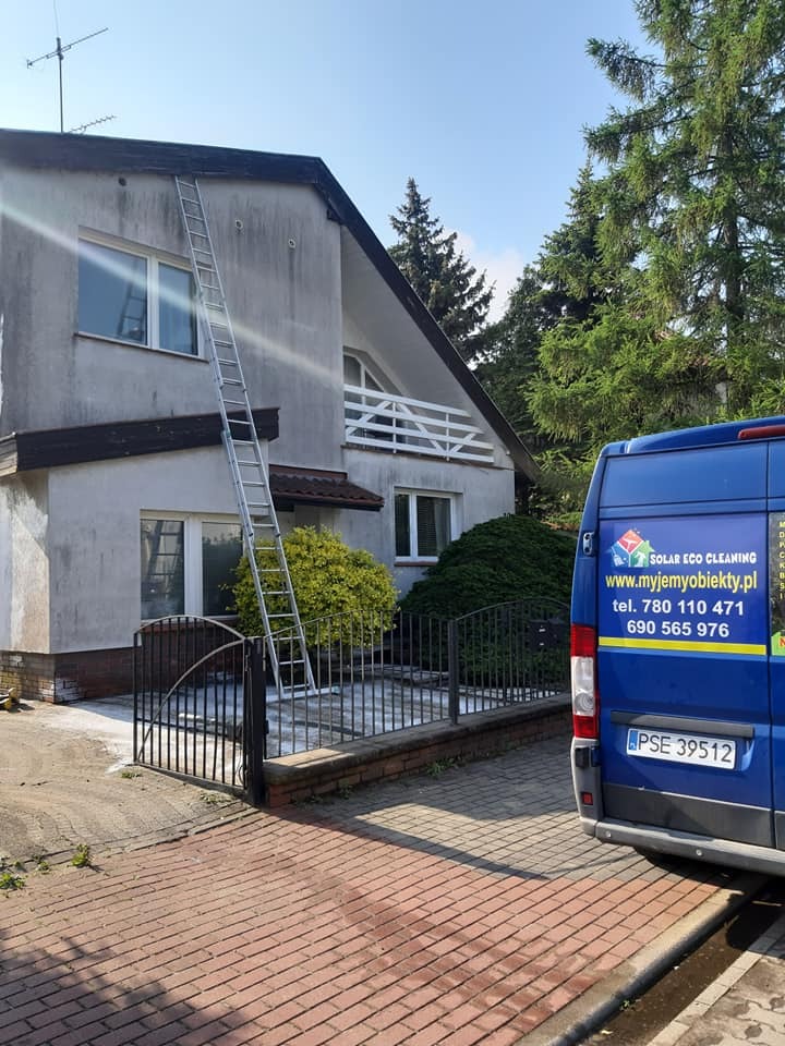 Dwupiętrowy dom z szarą elewacją przed czyszczeniem, z drabiną opartą o ścianę i samochodem firmy sprzątającej w tle, na tle zieleni.