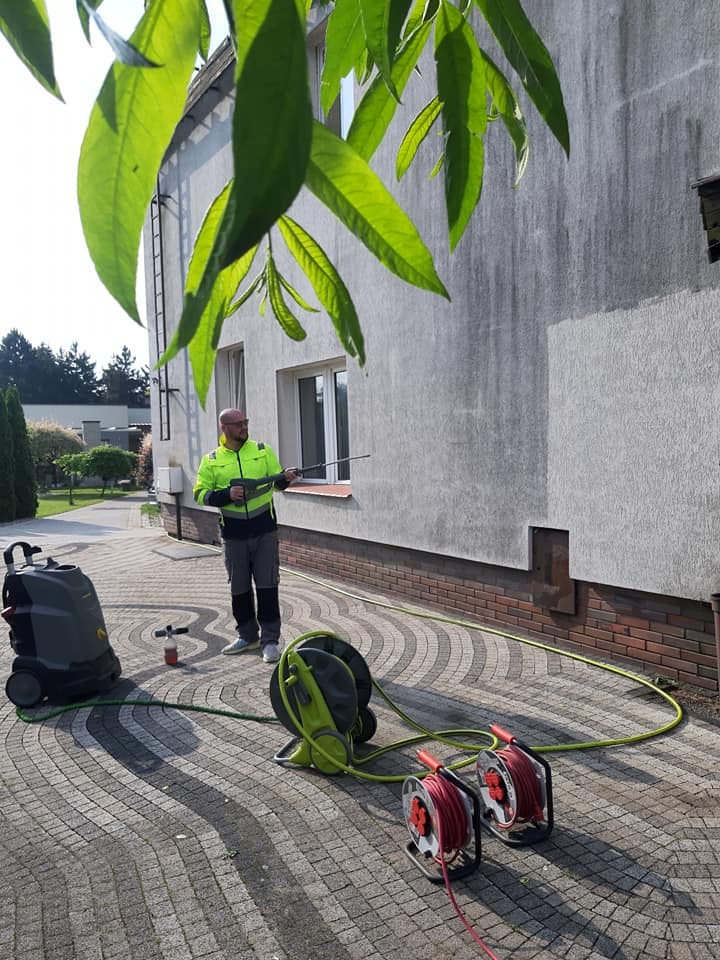 Pracownik w odblaskowej kurtce czyści elewację domu myjką ciśnieniową. Widoczny sprzęt, wąż i kostka brukowa. Ujęcie z dołu, z liśćmi drzewa w kadrze.