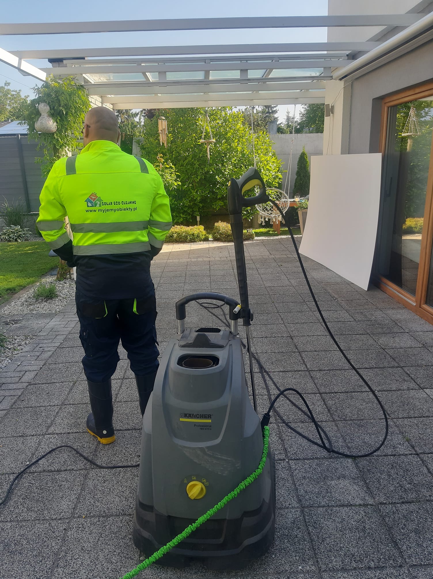 Pracownik w odblaskowej kurtce stoi tyłem obok myjki ciśnieniowej Kärcher na kostce brukowej. Widoczny fragment elewacji domu i zieleń w tle.