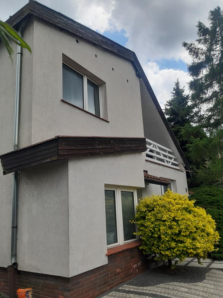 Jasna elewacja domu po czyszczeniu, widoczne okna, rynna, fragment dachu i kostki brukowej. Na pierwszym planie ozdobny krzew.