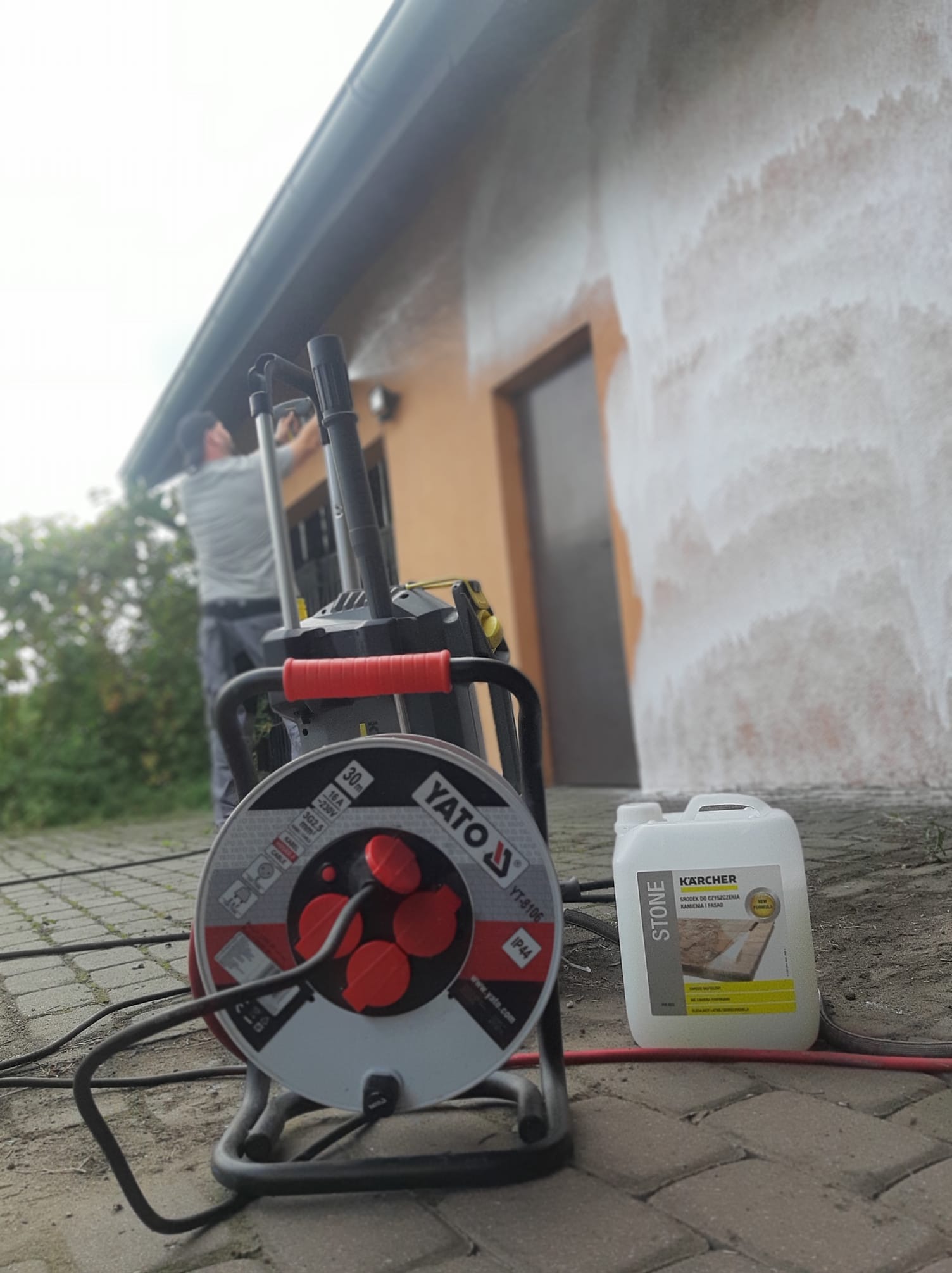 Czyszczenie elewacji domu myjką ciśnieniową; widoczny pracownik, maszyna Yato, środek czyszczący Kärcher Stone. Fragment ściany w trakcie mycia.