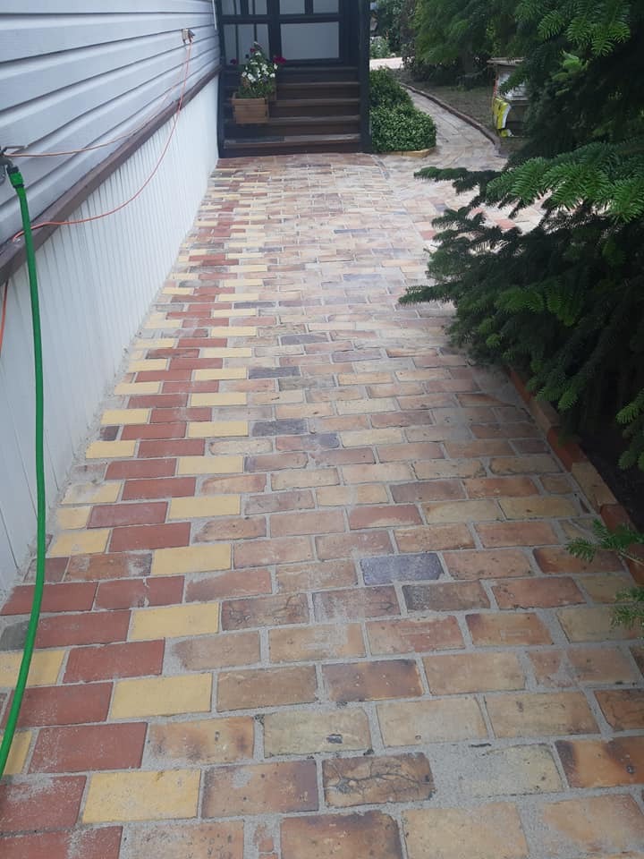 Ceglana ścieżka prowadząca do domu, z zielonym wężem ogrodowym i roślinnością po bokach. Widoczne schody wejściowe i fragment elewacji.