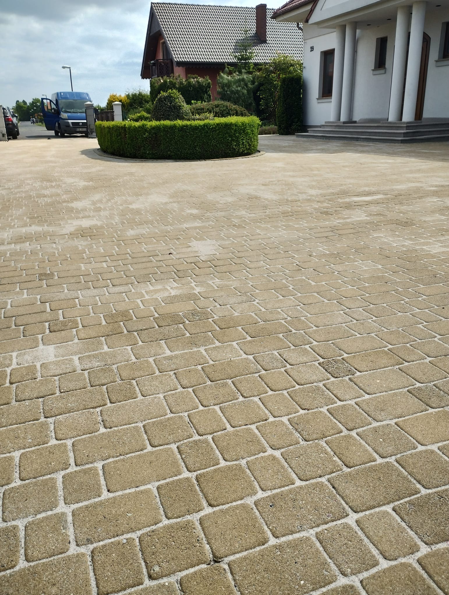 Jasna kostka brukowa na podjeździe, widok z góry. W tle dom z białymi kolumnami i zadbany żywopłot. Na podjeździe stoi bus z otwartymi drzwiami.