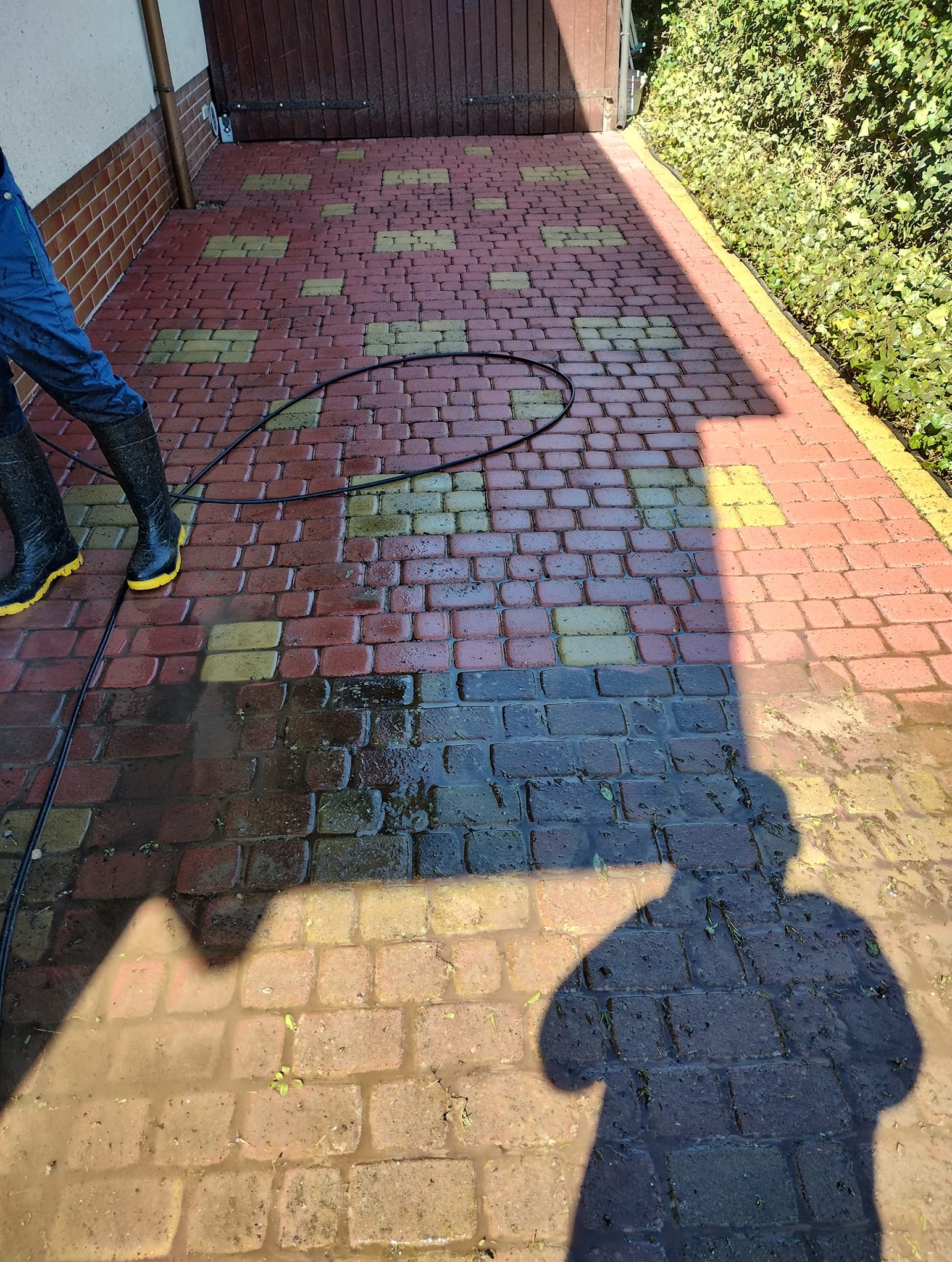 Mycie kostki brukowej z użyciem myjki ciśnieniowej. Widoczne różnice w kolorze czystej i brudnej kostki. Cień osoby obsługującej myjkę.