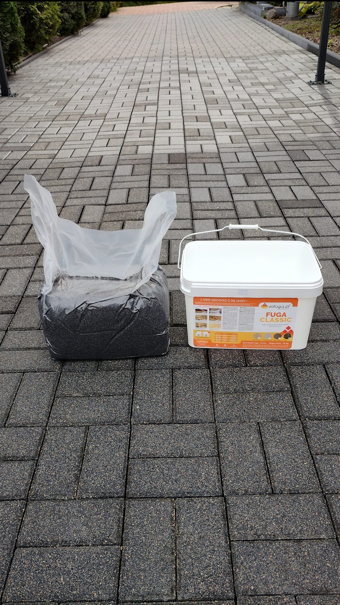 Szara kostka brukowa, obok worek z czarną fugą i białe wiaderko z napisem 'Fuga Classic' na tle wybrukowanej ścieżki.